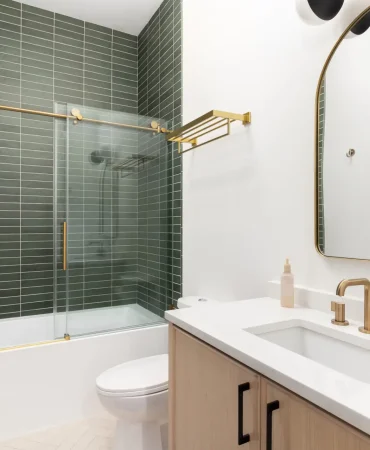 A bathroom with a wood vanity cabinet, gold hardware and faucet, and a shower with green subway tiles.