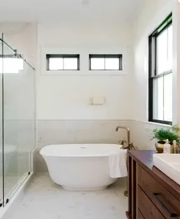 A modern bathroom featuring a sleek freestanding tub, glass-enclosed shower, wooden vanity with round mirror, brass fixtures, and bright natural lighting.